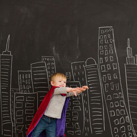 Cute toddler boy (2-3) standing against blackboard with city skyline drawn on it. Photo : Jessica Peterson