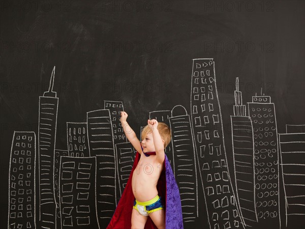 Cute toddler boy (2-3) standing against blackboard with city skyline drawn on it. Photo : Jessica Peterson