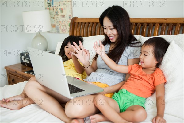 Mother with daughters (2-3, 4-5) sitting on bed, using laptop, waving. Photo : Jessica Peterson