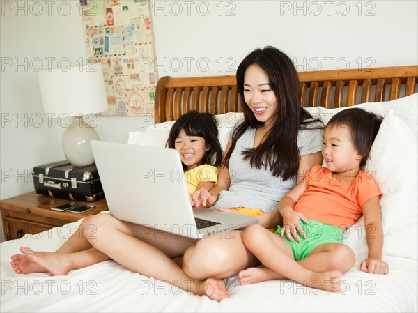 Mother with daughters (2-3, 4-5) sitting on bed, using laptop. Photo : Jessica Peterson