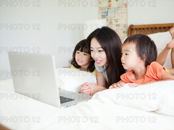 Mother with daughters (2-3, 4-5) lying down in bed, using laptop. Photo : Jessica Peterson