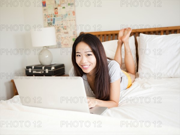 Young woman lying down in bed, using laptop. Photo : Jessica Peterson