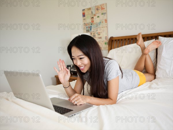 Young woman lying down in bed, using laptop, waving. Photo : Jessica Peterson