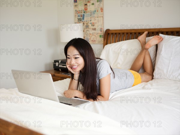 Young woman lying down in bed, using laptop. Photo : Jessica Peterson