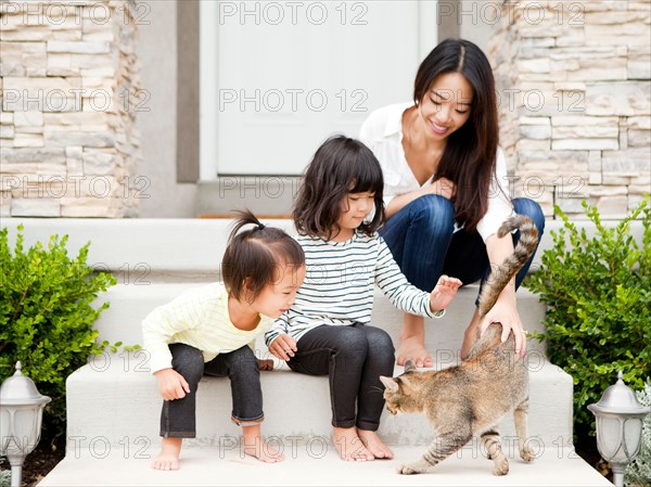 Mother sitting on doorsteps with two daughters (2-3, 4-5) and playing with cat. Photo : Jessica Peterson