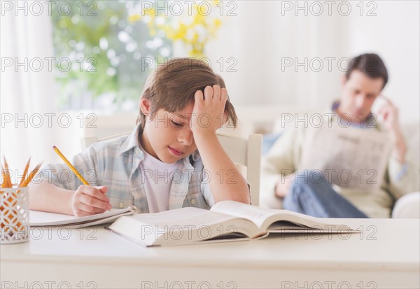 Boy (10-11 years) doing homework at table with father sitting in background. 
Photo : Daniel Grill