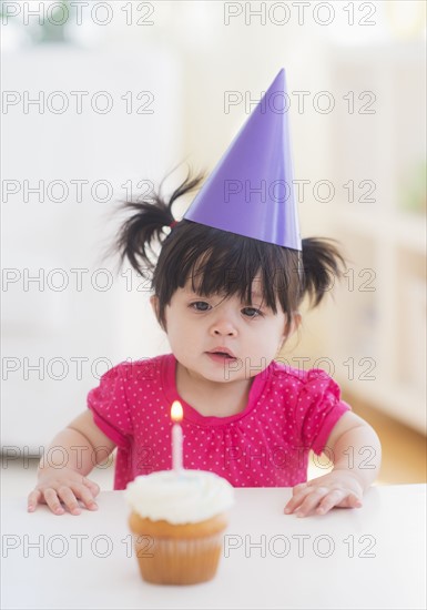 Portrait of baby girl (12-17 months) in party hat with cupcake. 
Photo : Daniel Grill