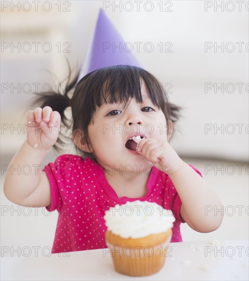 Portrait of baby girl (12-17 months) in party hat eating cupcake. 
Photo : Daniel Grill