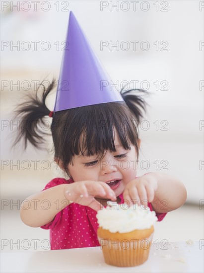 Portrait of baby girl (12-17 months) in party hat eating cupcake. 
Photo : Daniel Grill