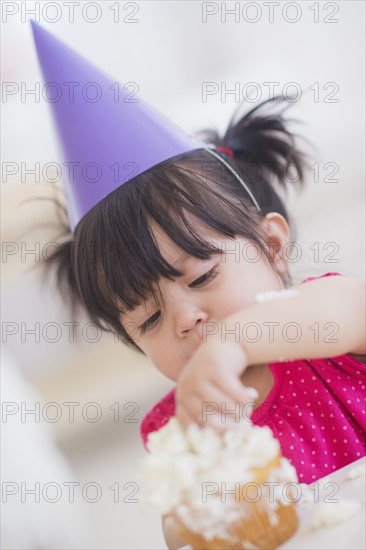 Portrait of baby girl (12-17 months) in party hat eating cupcake. 
Photo : Daniel Grill