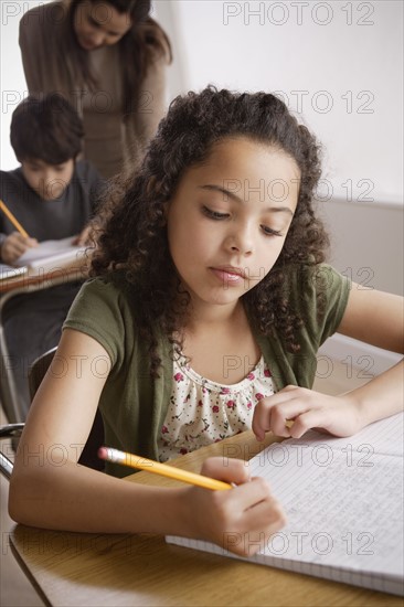 Schoolgirl (10-11) holding pencil with boy (12-13) and teacher in background. 
Photo: Rob Lewine