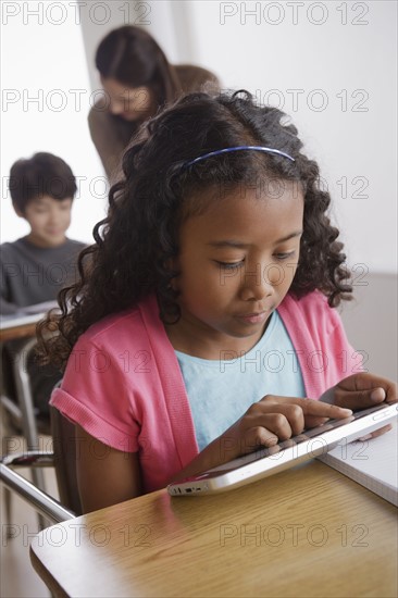 Schoolgirl (10-11) holding digital tablet with boy (12-13) and teacher in background. 
Photo : Rob Lewine