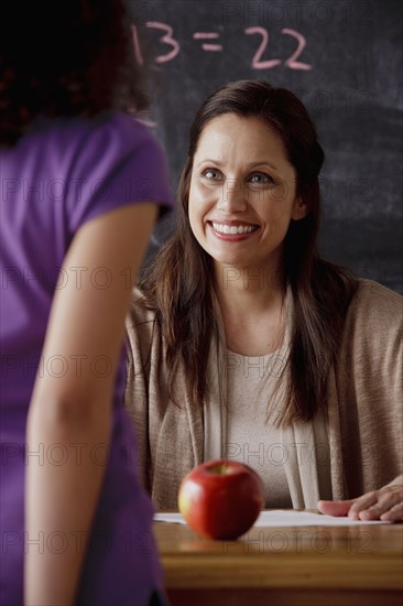 Smiling teacher with apple on deck schoolgirl (10-11) in foreground. 
Photo : Rob Lewine