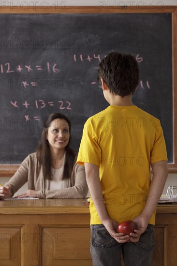 Schoolboy (12-13) holding apple behind back with teacher in background. 
Photo : Rob Lewine