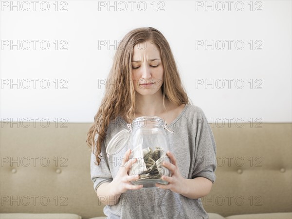 Young woman looking into savings jar with unhappy face expression . 
Photo : Jessica Peterson