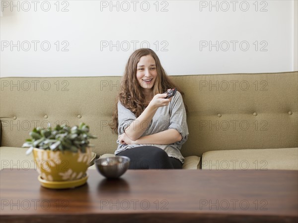 Cheerful young woman sitting on sofa with remote control. 
Photo : Jessica Peterson