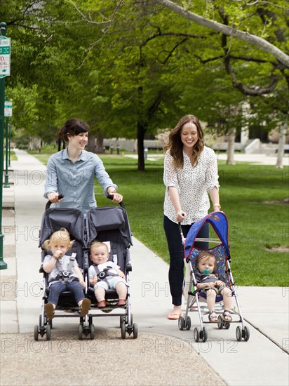 USA, Utah, Salt Lake City, Two young mothers walking with two baby boys (6-11 months) and toddler girl (2-3) in push prams. 
Photo : Jessica Peterson