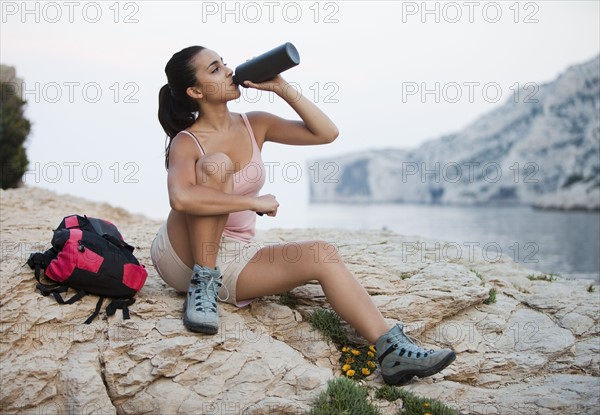 France, Marseille, Woman with backpack drinking water on rocky beach. Photo : Mike Kemp