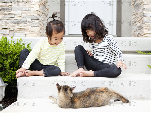 Sisters (2-3, 4-5) sitting in front of house with cat. Photo : Jessica Peterson