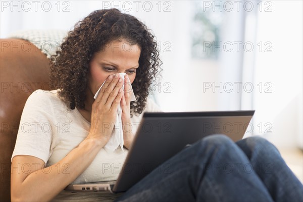 Portrait of woman sitting with laptop and blowing nose.