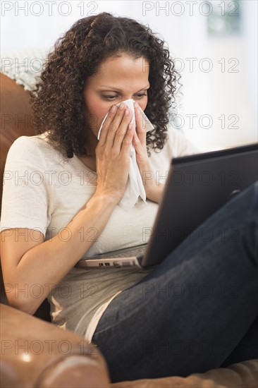 Portrait of woman sitting with laptop and blowing nose.