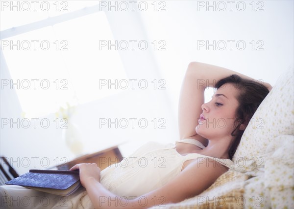 Woman in white chemise laying in bed and holding book. Photo : Daniel Grill