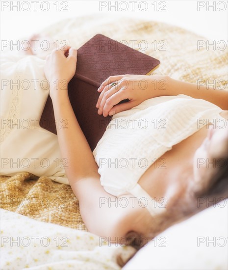 Woman in white chemise laying in bed and holding book. Photo : Daniel Grill