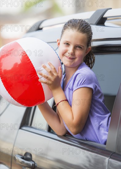 Young girl (8-9) with beach ball peeking through car window . Photo : Daniel Grill