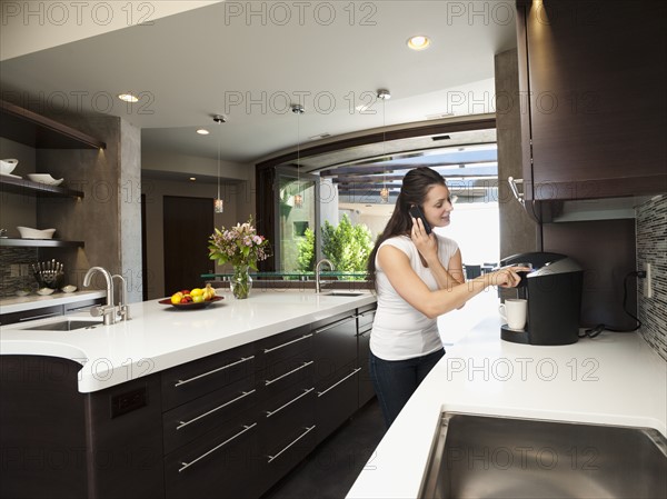 Young woman making morning coffee while using mobile phone.