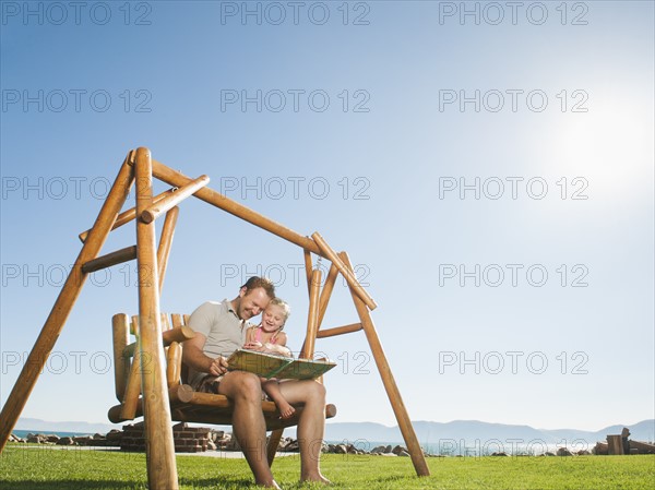 Father with daughter (4-5) on swing reading book.