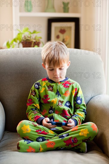 Boy in his pijamas sitting on armchair and playing video game.. Photo : Jessica Peterson