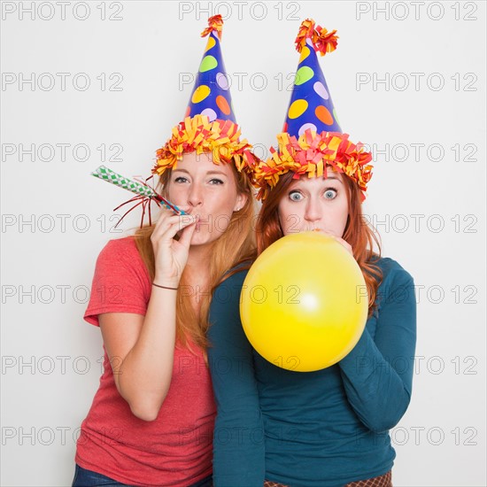 Studio Shot, Two women wearing party hats and one blowing up yellow balloon. Photo : Jessica Peterson