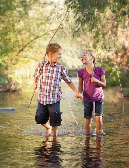 Little boy (6-7) and little girl (4-5) holding hands and walking together in small stream holding wooden stick fishing poles
