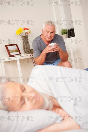 Senior man with coffee cup looking at sleeping wife. Photo : Rob Lewine