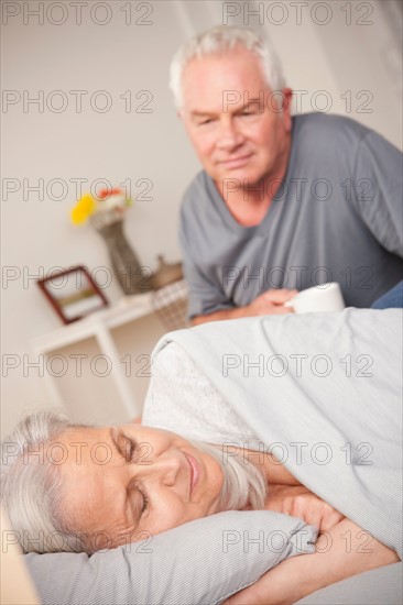 Senior man with coffee cup looking at sleeping wife. Photo : Rob Lewine