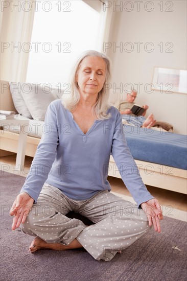 Senior woman practicing yoga with husband reading in background. Photo : Rob Lewine