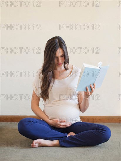 Pregnant mid adult woman sitting with crossed legs and holding book. Photo : Jessica Peterson