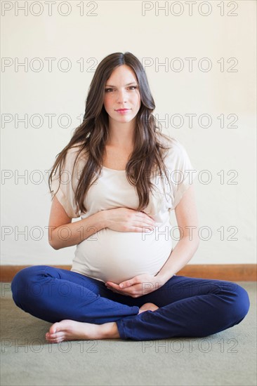 Portrait of pregnant mid adult woman sitting with crossed legs touching her abdomen. Photo: Jessica Peterson