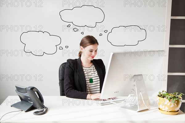 Studio shot of young woman working at desk with thought bubbles around her head. Photo: Jessica Peterson