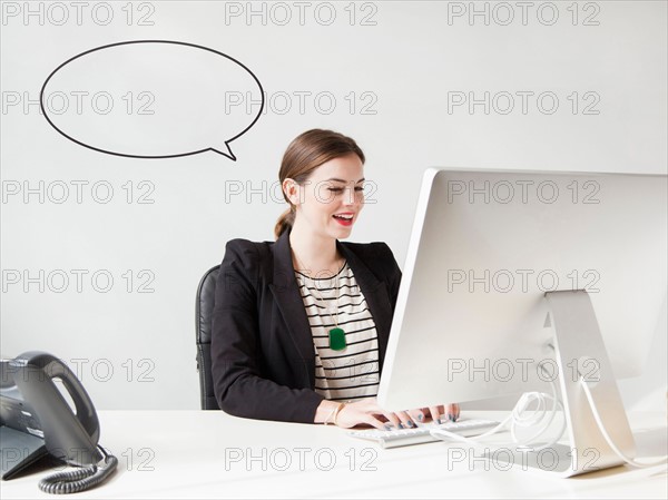 Studio shot of young woman working on computer with speech bubble next to her head. Photo: Jessica Peterson