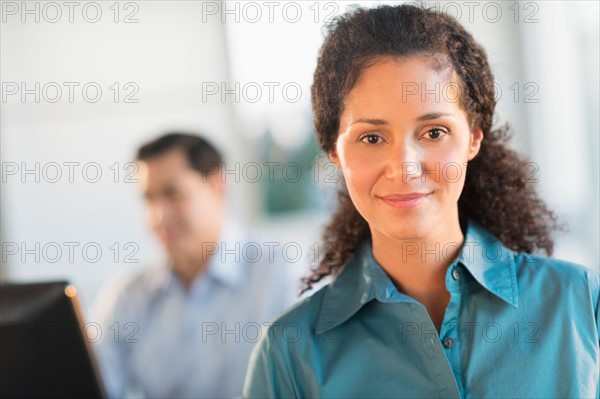 Portrait of businesswoman in office with male executive working in background.