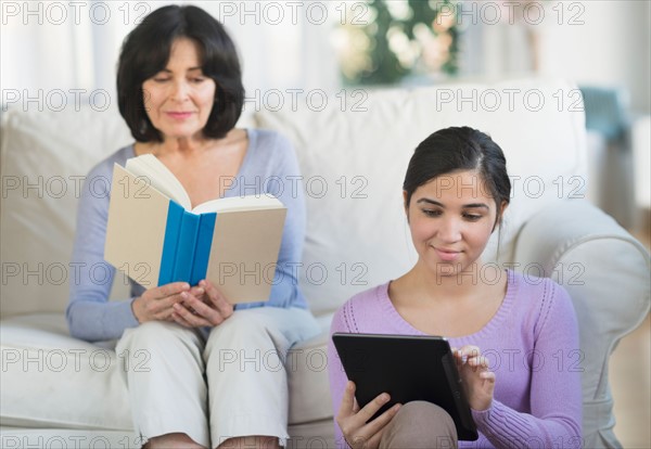 Grandmother reading book and granddaughter (16-17) sitting with digital tablet.