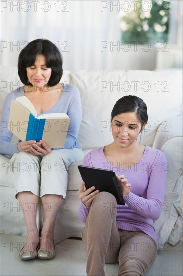 Grandmother reading book and granddaughter (16-17) sitting with digital tablet.