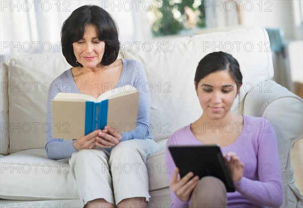 Grandmother reading book and granddaughter (16-17) sitting with digital tablet.