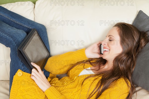 Woman lying on sofa with digital tablet and talking via cell phone