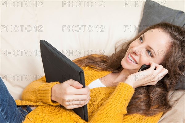 Woman lying on sofa with digital tablet and talking via cell phone