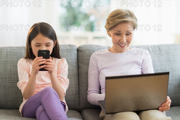 Granddaughter (8-9) and grandmother sitting on sofa and using laptop and cell phone.