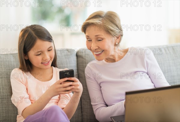 Granddaughter (8-9) and grandmother sitting on sofa and using laptop and cell phone.