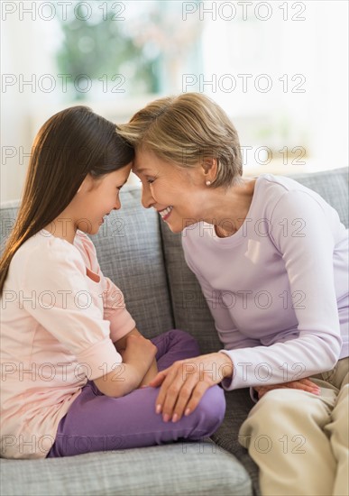 Granddaughter (8-9) and grandmother sitting on sofa and touching with heads.