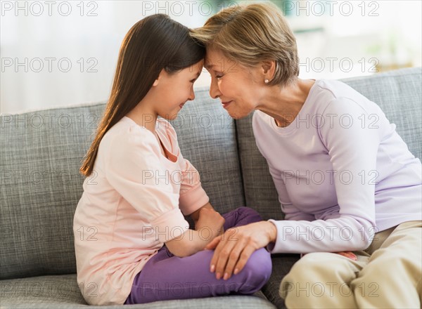 Granddaughter (8-9) and grandmother sitting on sofa and touching with heads.